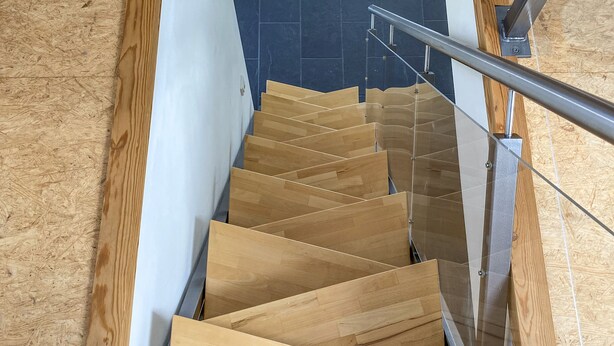 Eine Holztreppe mit Glastrennwand, die zu einem dunklen Fliesenboden führt.