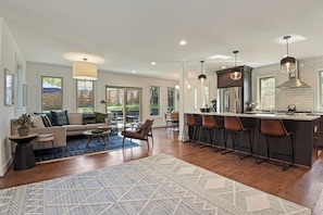 A spacious living area with a sofa, coffee table, and a patterned rug. A kitchen area with a bar and stools is visible. - The Crestwood House - Family Friendly Retreat with Private Backyard - DCA  (Alexandria)