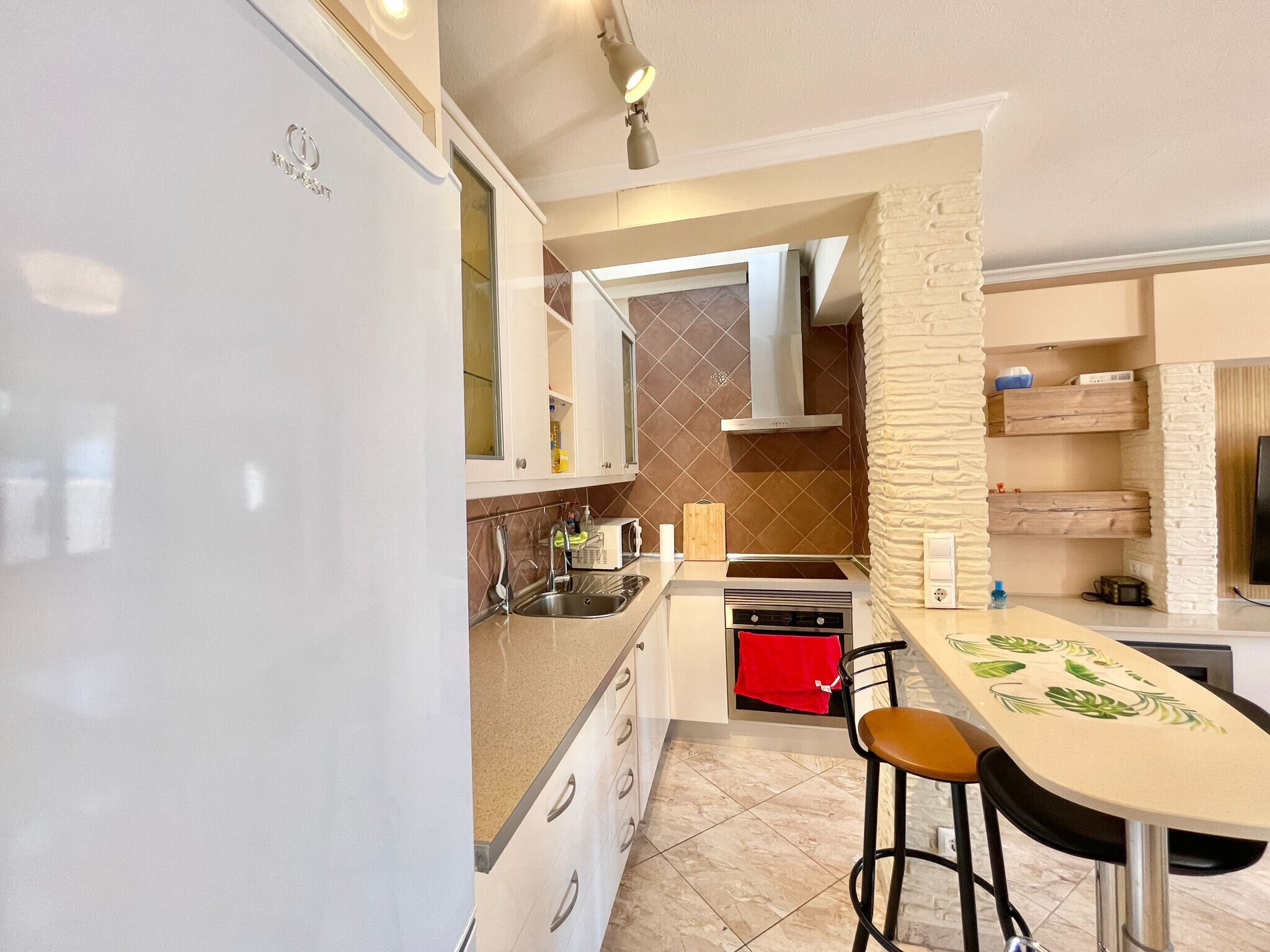 A modern kitchen with a white refrigerator, stainless steel sink, and a red oven. There are two stools at a table with a green leaf pattern.