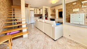Een moderne keuken met witte kasten, een inbouwoven en een zithoek met een witte bank en een houten tafel.