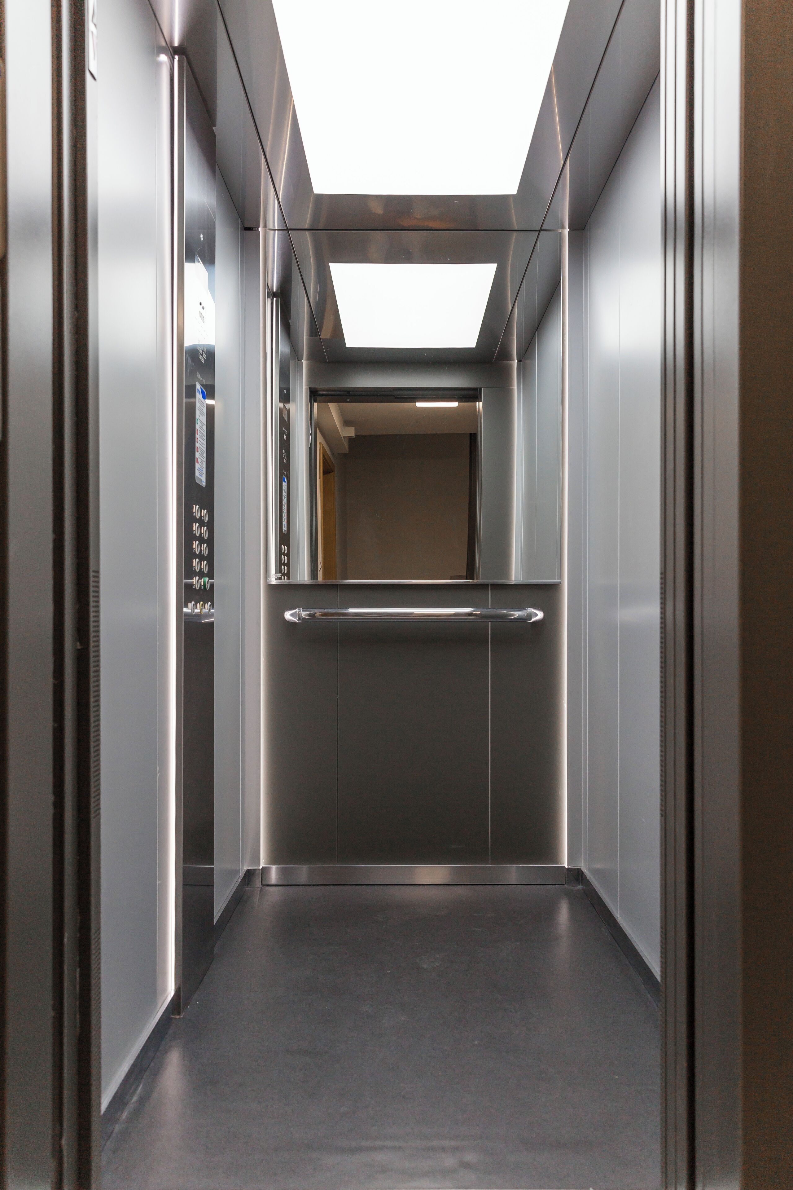 A modern elevator with a metallic interior, a handrail, and a control panel on the left.