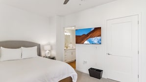 Ein Schlafzimmer mit einem Bett, einem Nachttisch, einem an der Wand befestigten Fernseher, einer Tür und einer kleinen Mülltonne.