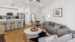 Eine moderne Küche mit weißen Schränken, einer zentralen Kochinsel mit Barhocker, einem Deckenventilator und einem Essbereich mit grauem Sofa und rundem Couchtisch.