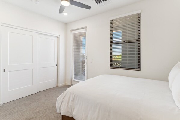 Une chambre à coucher avec un lit, un ventilateur de plafond, une porte et une fenêtre avec des stores.