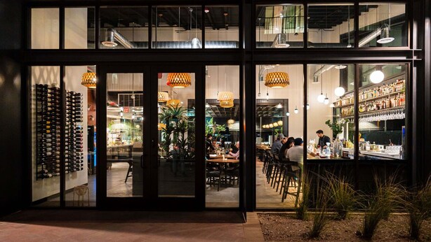 A restaurant with a modern interior, featuring a bar area with shelves of bottles and a seating area with tables and chairs.
