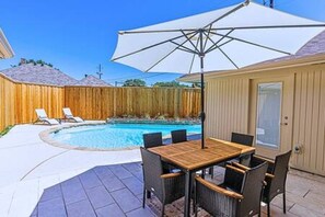 Un jardin avec une piscine, une terrasse en bois, une table de patio couverte et des chaises longues.