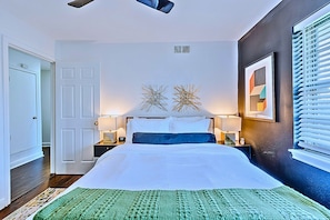 Habitación de hotel con cama, dos mesitas de noche, una lámpara de noche, un tapete de área verde, una obra de arte en la pared y una ventana con persianas.
