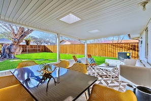 Un patio exterior con mesa y sillas, una parrilla y vistas a un jardín trasero con cerca de madera y un árbol.