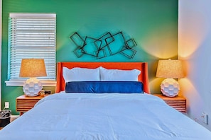 A bed with white bedding, a blue accent pillow, and a geometric wall art piece above it.