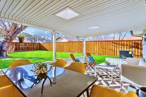 A covered patio with a table and chairs, a patterned floor, and a view of a backyard with a fence and greenery.