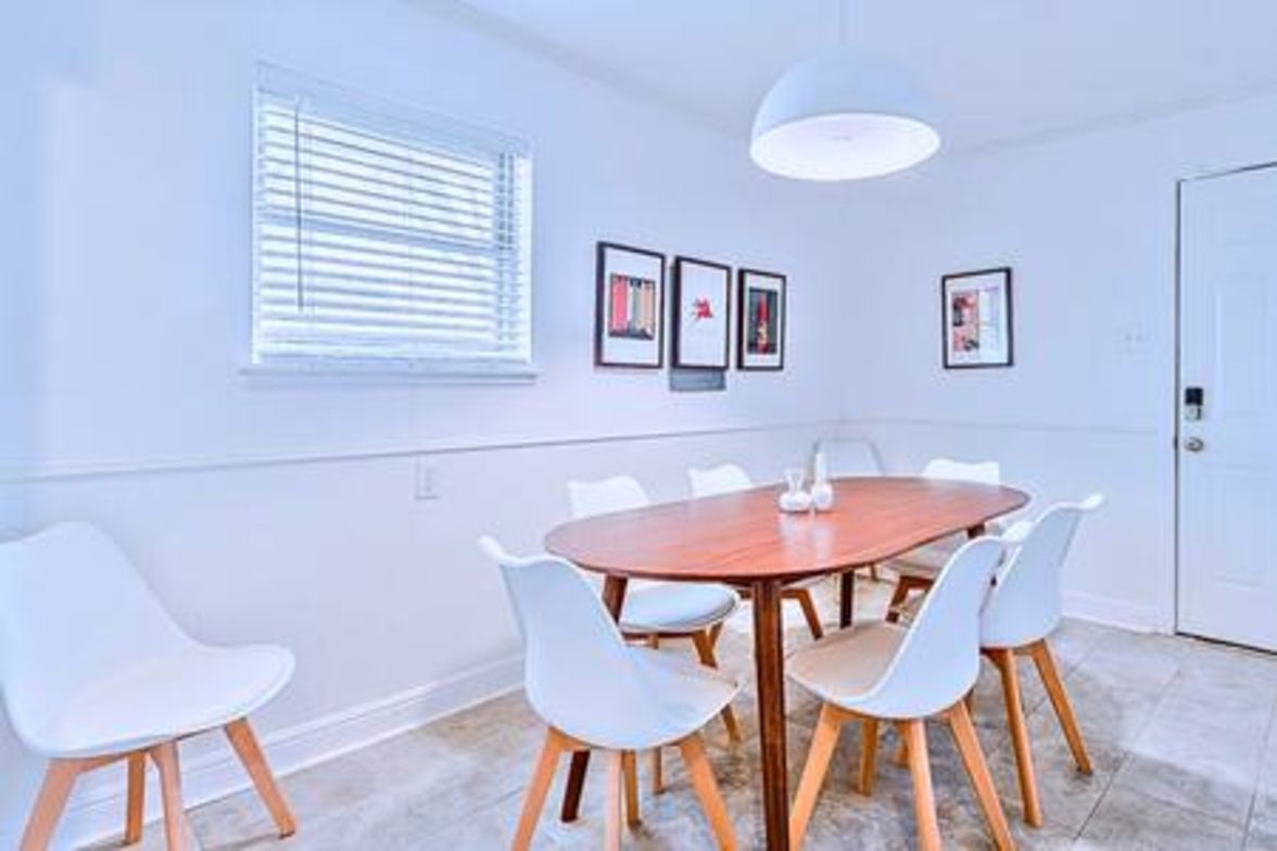 A room with a round wooden table, white chairs, a window with blinds, and framed pictures on the wall.