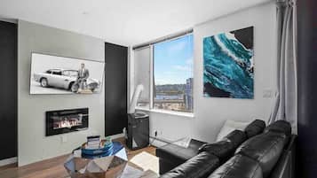 A modern living room with a black leather couch, a fireplace, a large window with a city view, and artwork on the walls.