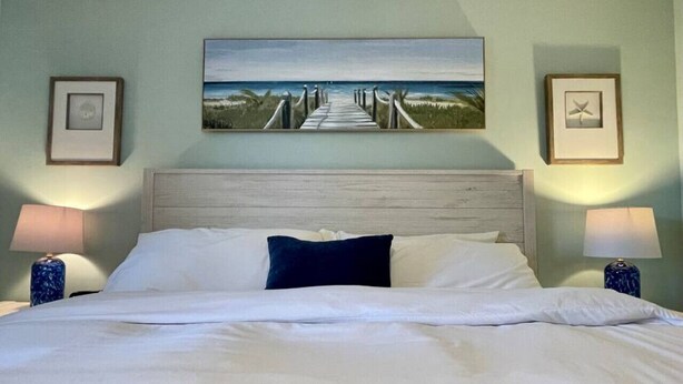 A neatly made bed with white linens and a dark blue pillow, flanked by two bedside lamps with blue bases. Above the bed, there is a framed painting of a beach pathway. - Sunrise & Sunset views, Pool, 1/2 mile to beach! (Jupiter)