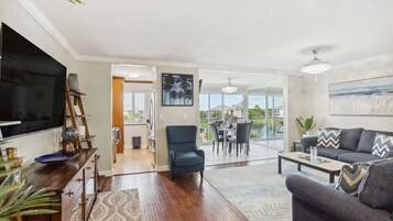 A spacious living room with a large TV, a blue armchair, a gray sofa, and a dining area with a table and chairs.