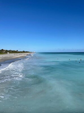 Une plage avec une eau turquoise et limpide, un rivage sablonneux et quelques bâtiments en arrière-plan.