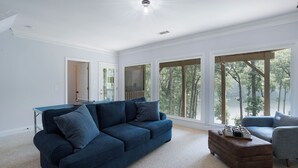 Ein geräumiges Wohnzimmer mit einem blauen Sofa, einem Couchtisch und großen Fenstern mit Blick auf Bäume.