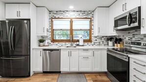 A modern kitchen with white cabinets, stainless steel appliances, a window with blinds, and a gray rug on the floor. - Fire Pit, Fully Stocked, Family-Friendly Oasis (Wichita)