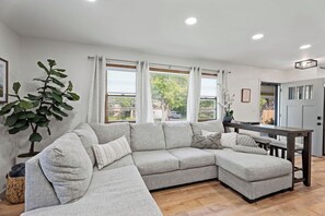 A modern living room with a sectional sofa, a dining table, and a large window with curtains. - Fire Pit, Fully Stocked, Family-Friendly Oasis (Wichita)