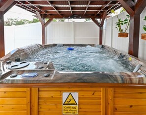 Banheira de hidromassagem com placa de aviso sobre pisos escorregadios quando molhados, cercada por estruturas de madeira e um cercado branco.