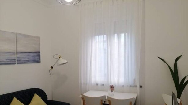 Een kleine, verzorgde kamer met een zwarte bank, een witte eettafel met twee stoelen, en een losse stoel. Op de tafel staat een lamp, rechts een plant en aan de muur een schilderij.