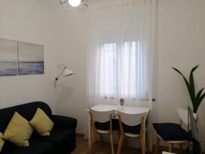 Ett litet, välordnat rum med en svart soffa, ett vitt matbord med två stolar och en ensam stol. På bordet står en lampa, till höger står en växt och på väggen hänger en tavla.