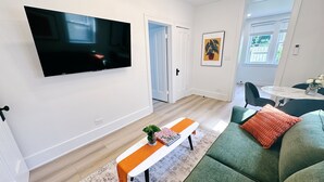 Moderni olohuone, jossa seinälle on asennettu taulutelevisio, on vihreä sohva, valkoinen sohvapöytä, jonka päällä on kirja ja maljakko, sekä kuviollinen matto.