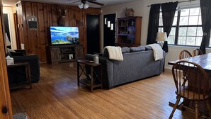 A cozy living room with a wooden floor, a gray sofa, a TV, a ceiling fan, and a dining area with a wooden table and chairs. - Dragons Lair - motorcycle friendly, all paved roads, lockable garage, no incline (Robbinsville)