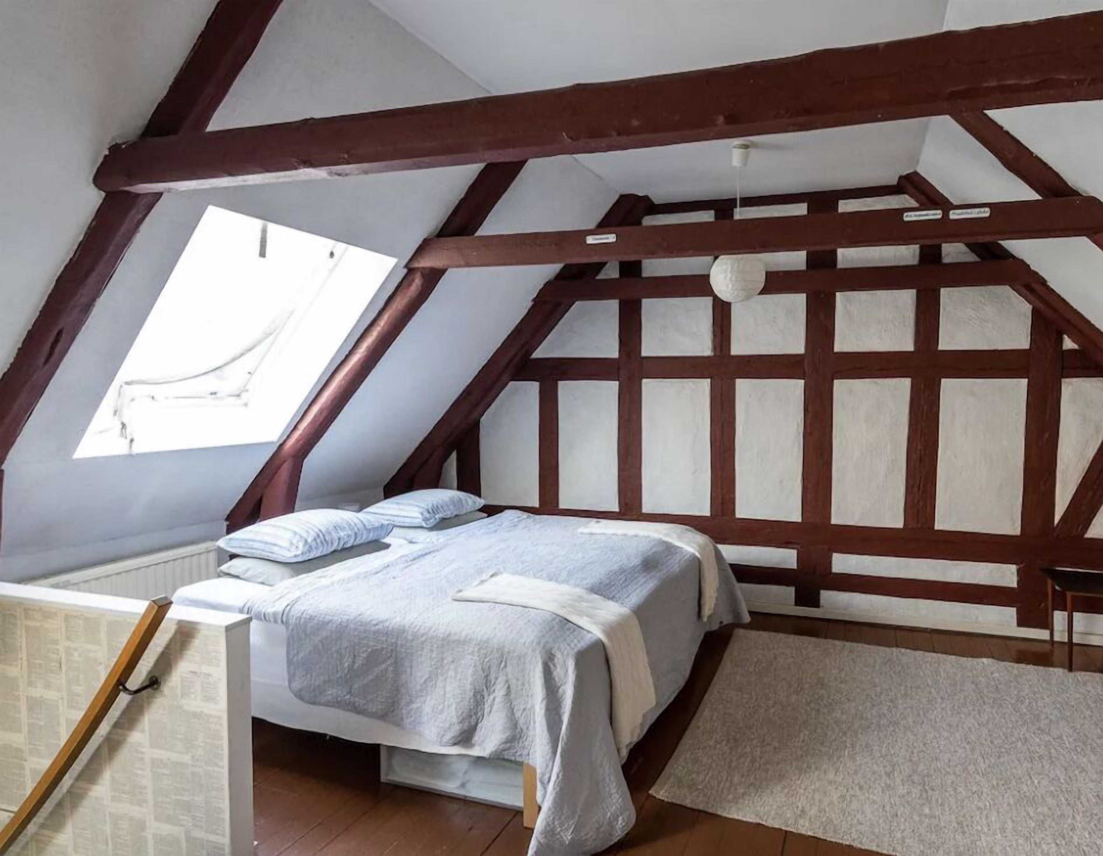 Ein Schlafzimmer mit schrägverdachter Decke, freiliegenden Holzbalken, einem Bett mit weißer und grauer Bettdecke und einem kleinen Fenster.