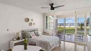 A bedroom with a bed, bedside table, lamp, and a view of the outdoors through a sliding glass door. - Beachfront Petite Paradise unit at Pelican Reef 103 (Rincon)