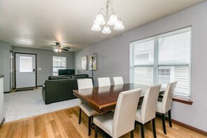 A dining area with a wooden table and white chairs, a ceiling fan, and a large window with blinds. - Stadium House - Next to Kernels/Roughriders Arena (Cedar Rapids)