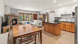 A modern kitchen with wooden cabinets, a central island, a stone fireplace, and a dining area with a wooden table and chairs.