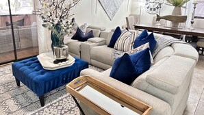 Ein Wohnzimmer mit einem beigen Sofa, blau-weißen Kissen, einem blauen Hocker, einer Dekovase und einer Wanduhr.