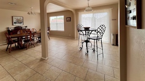A spacious dining area with a wooden table and chairs, a kitchen in the background, and a large window with blinds.