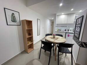 A compact kitchen with white cabinets, a dining table with four chairs, and a small wooden shelf unit.