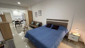 A modern hotel room with a large bed, a bedside table with a lamp, a small table with chairs, a sofa, and a kitchenette in the background. - Studio In San Telmo Paseo De La Cisterna 323 (Buenos Aires)