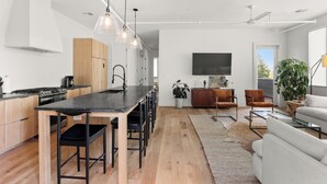 A modern kitchen with a central island, wooden cabinets, and a dining area with bar stools. There is a flat-screen TV mounted on the wall, a large potted plant, and a sofa in the living area. - 5BR City Loft - Atop Natural Wine Bar! Steps to Downtown & Magazine!  (New Orleans)