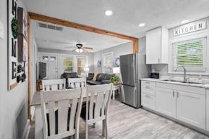 A modern kitchen with white cabinetry, a stainless steel refrigerator, and a dining area with white chairs and a wooden table. - Cottage on Walton (Indianapolis)
