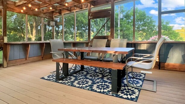 Eine Terrasse im Freien mit einem Holztisch und Stühlen, umgeben von großen Fenstern mit Blick auf Bäume und einen klaren Himmel.