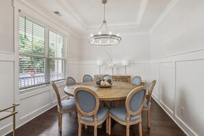 Un comedor con una mesa redonda de madera, seis sillas tapizadas, una araña y una ventana con persianas.