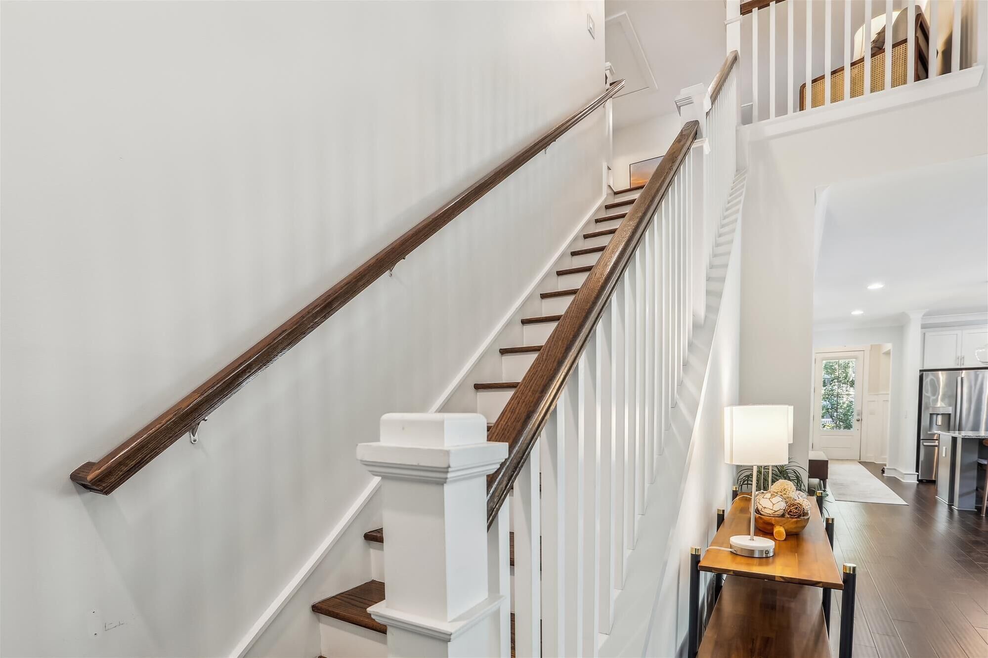 Una escalera con barandillas blancas y pasamanos de madera, que lleva a un rellano con una mesa y una lámpara.