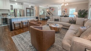 En rummelig stue med en stor beige sofa, et sofabord og et tæppe. Der er et køkkenområde med hvide skabe og rustfrie stålgre.