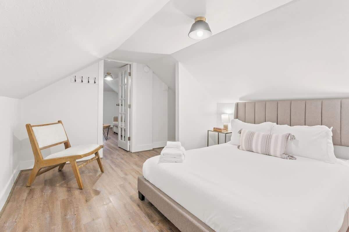 Quarto com cama, uma cadeira de madeira, mesa de cabeceira e porta que dá para outro espaço.