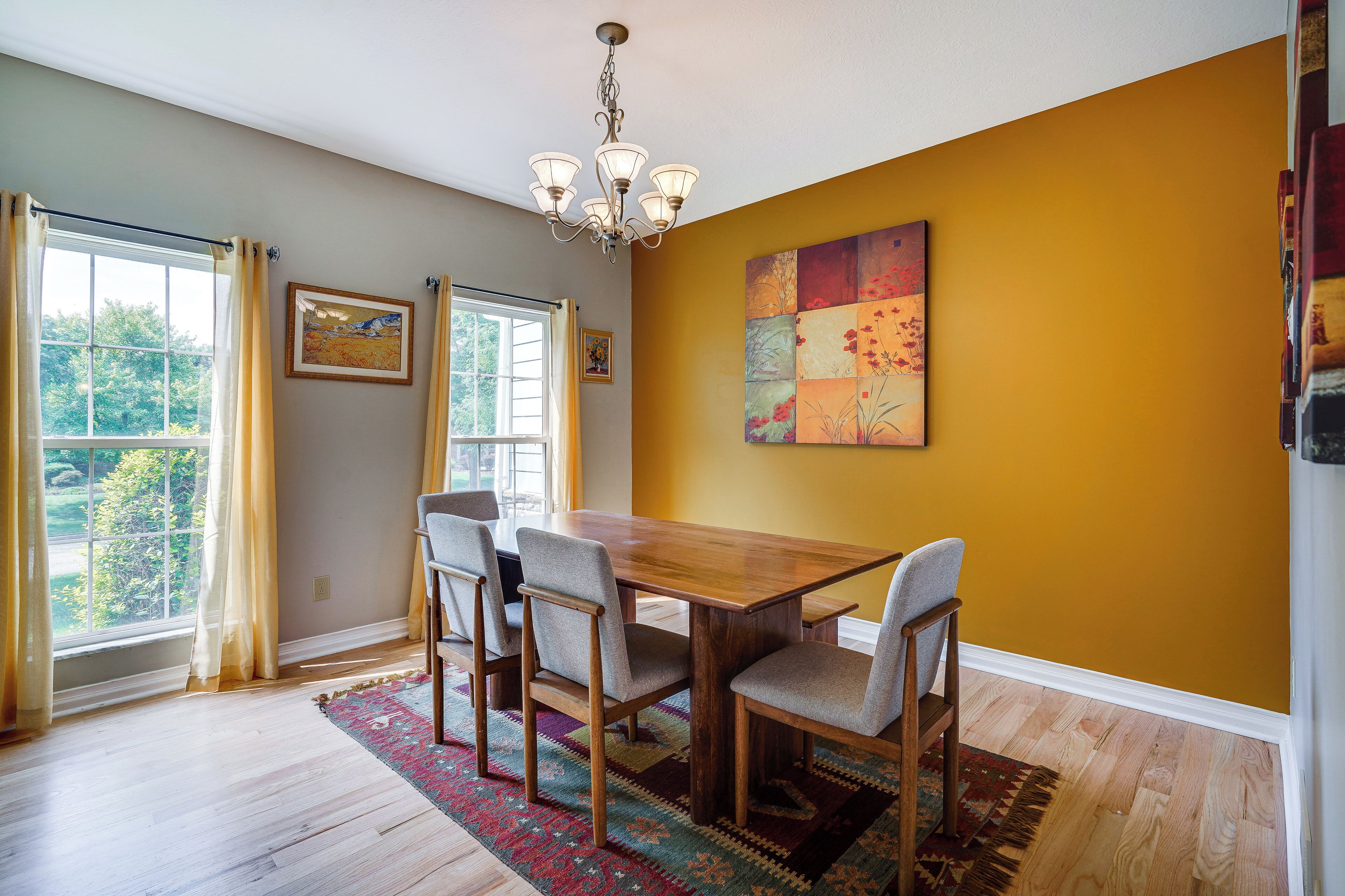 A dining area with a wooden table and four chairs, a chandelier, a colorful rug, and a large abstract painting on the wall.
