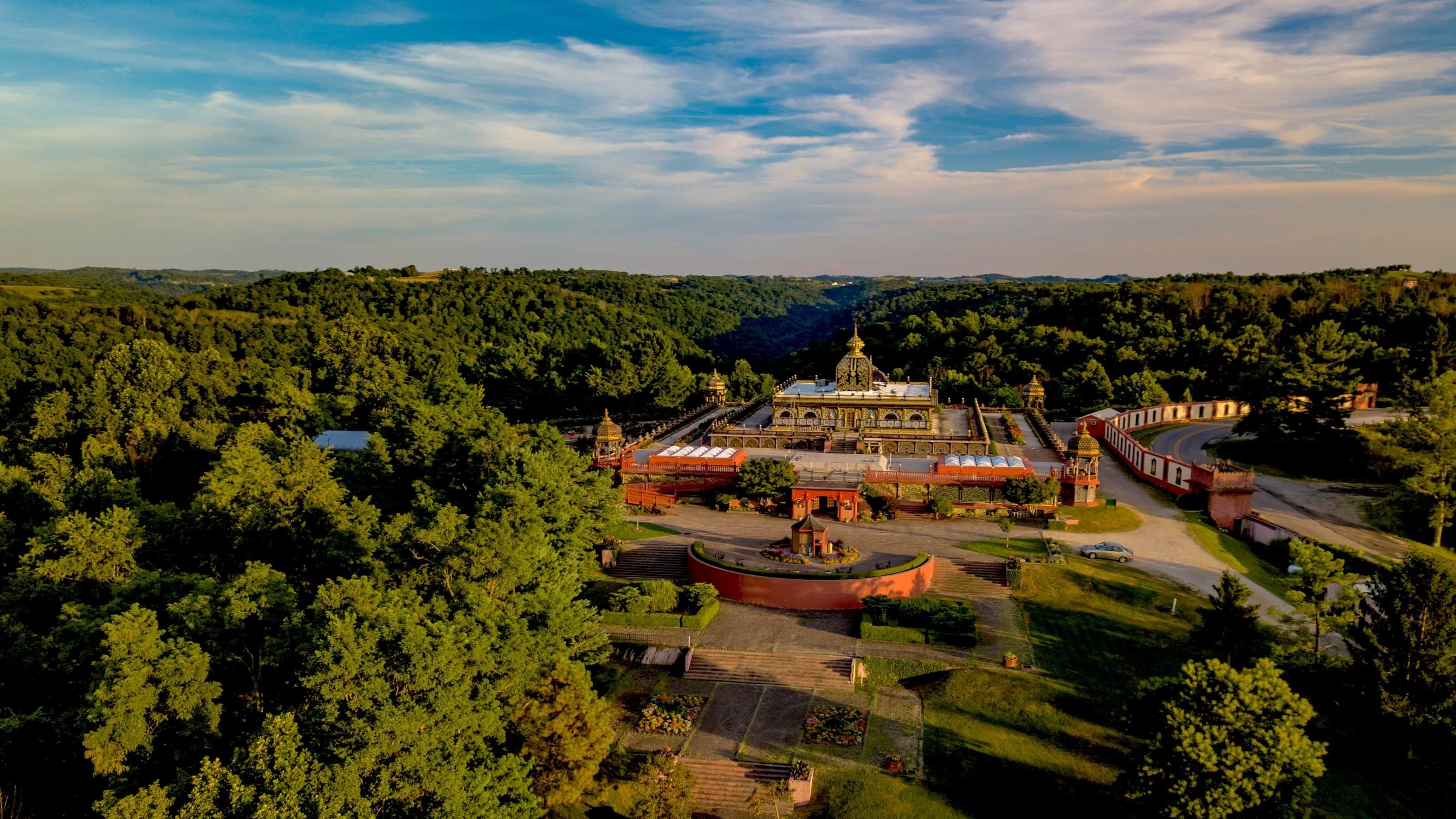 An aerial view of a historic building complex surrounded by dense forest, featuring a central domed structure, multiple smaller domes, and a symmetrical layout with pathways and gardens.