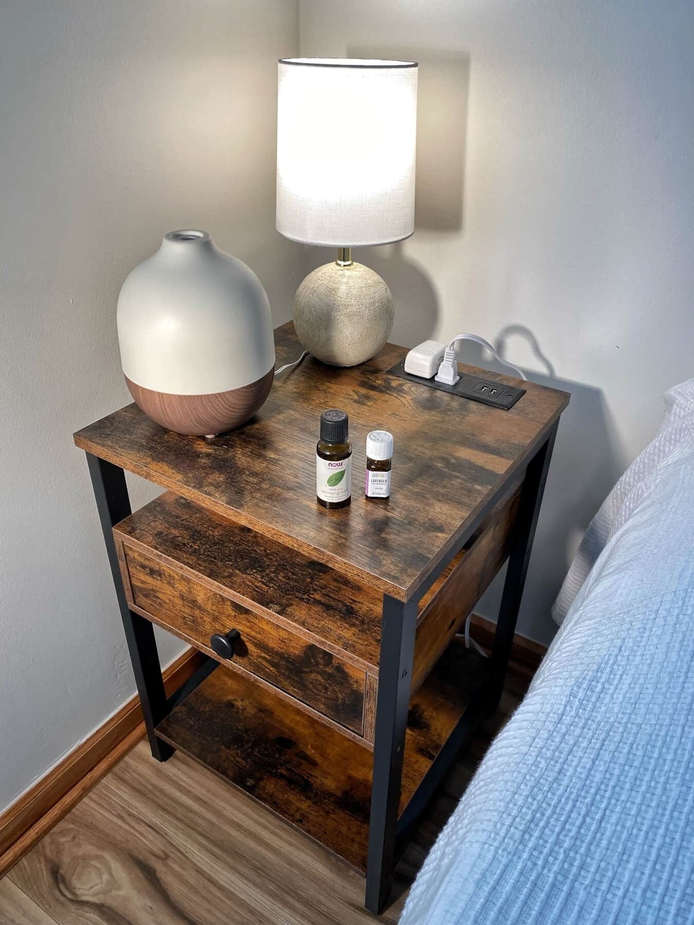 Une table de chevet en bois, sur laquelle se trouvent une lampe blanche, un vase et deux petites bouteilles.