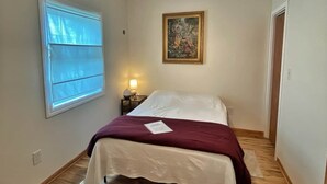 A bedroom with a bed, a ceiling fan, a window with blinds, a nightstand with a lamp, and a painting on the wall.