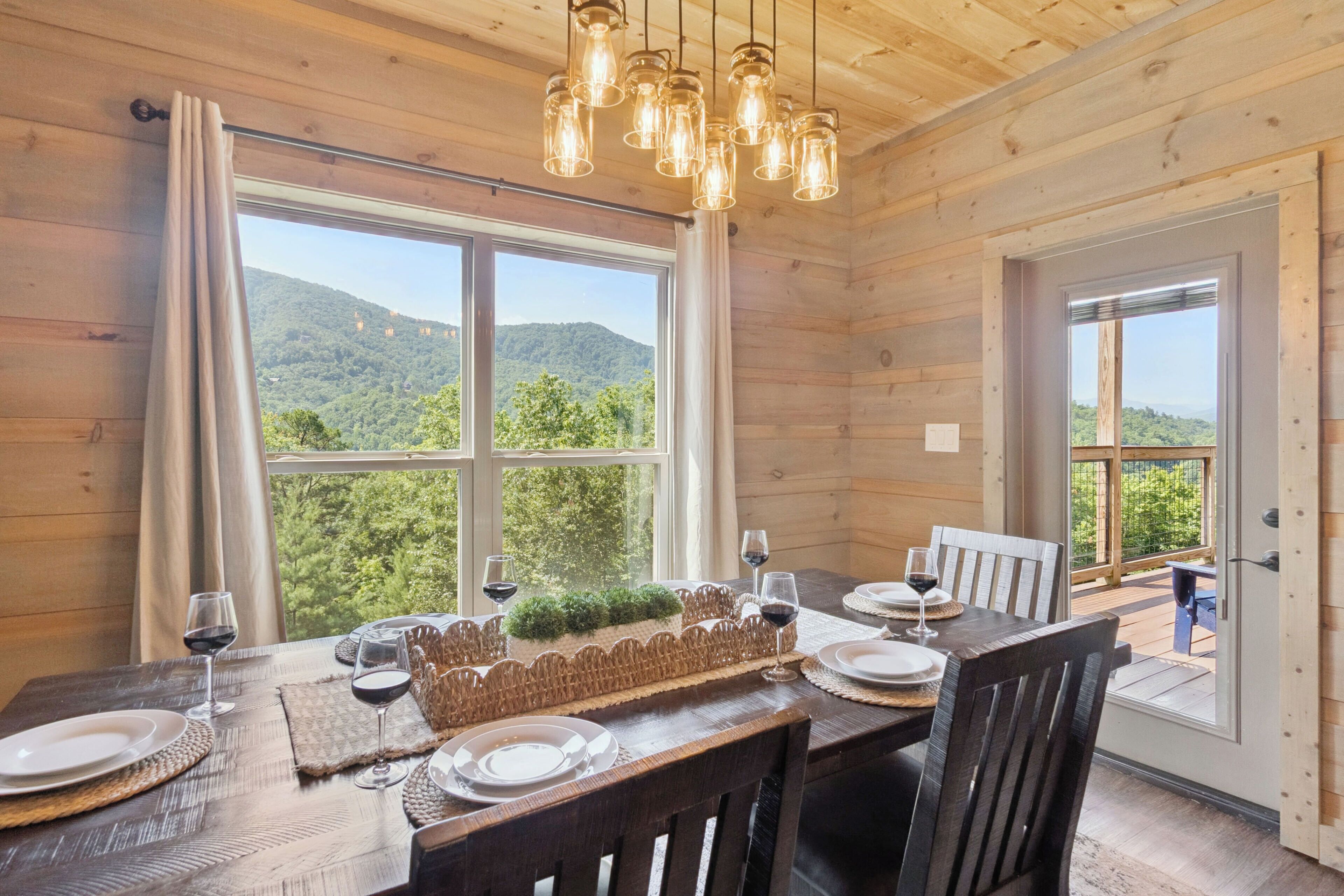 A wooden interior with a dining table set for two, two wine glasses, and a view of a forested mountain through large windows.