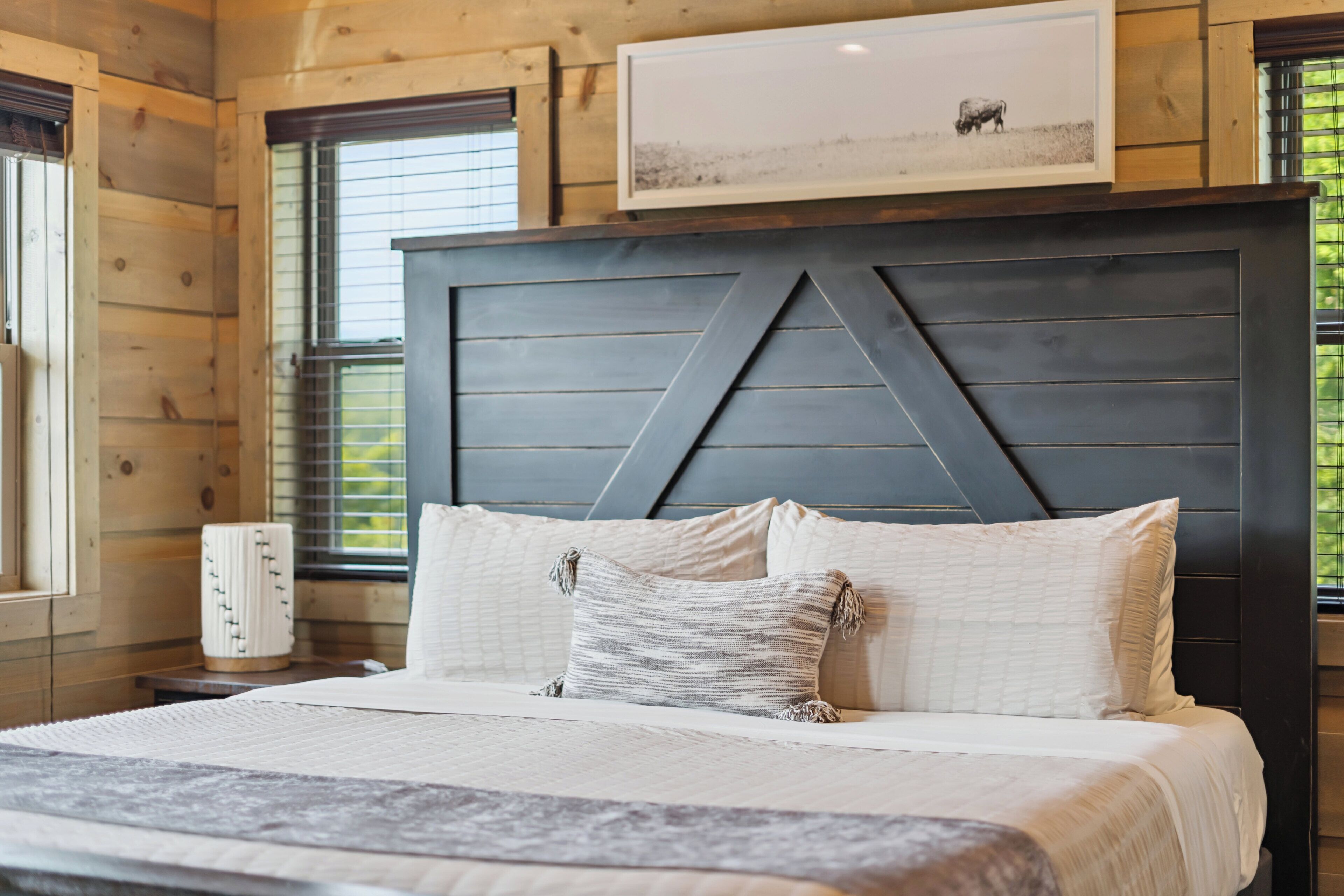 A bedroom with a large bed, a headboard with a geometric design, a framed picture above the bed, and a window with blinds.