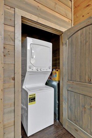 Pequeña lavandería compacta y blanca, con revestimiento de madera, un refrigerador pequeño y una botella de detergente.