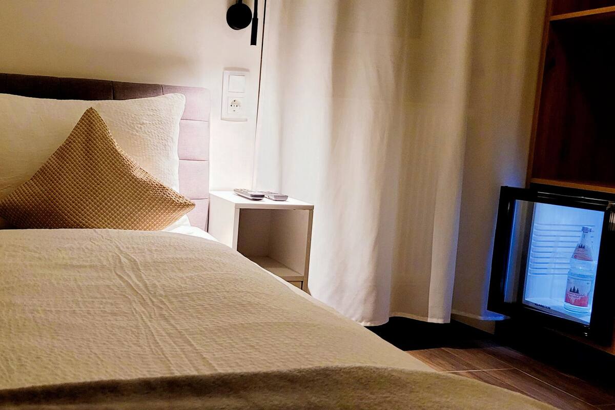 A bed with a wooden headboard, a white pillow, and a brown accent pillow. A small bedside table with a remote control and a bottle is visible.